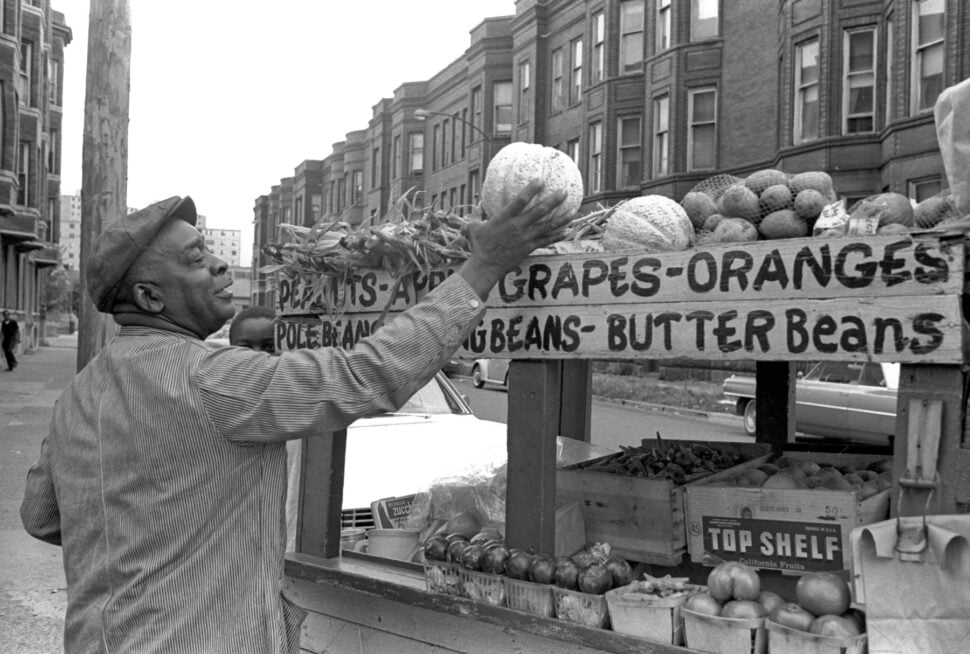 An elderly pushcart vendor reaches up to choose a melon for a customer, on a stop along his route in Bronzeville, a South Side neighborhood of Chicago, 1971