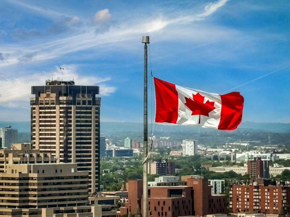A Canadian flag waves on a tall flagpole above a city skyline, with mid- and high-rise buildings below and a blue sky with wispy clouds in the background.