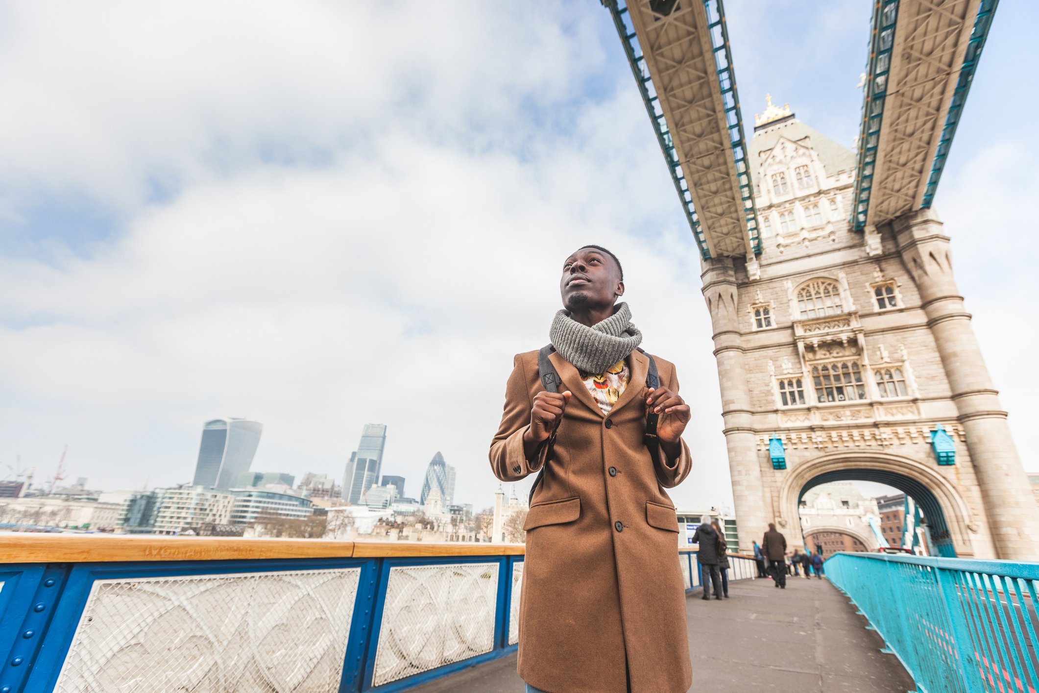 A person wearing a long brown coat and scarf stands on Tower Bridge in London, looking upward while holding backpack straps; the bridge structure frames the scene, with the city skyline and cloudy sky in the distance.
