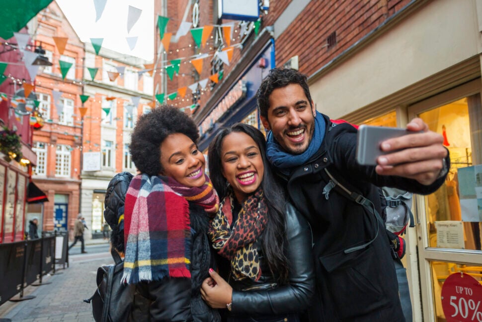 Three friends stand close together on a narrow city street decorated with green, white, and orange bunting, smiling and taking a selfie with a smartphone; shops and brick buildings line the street behind them in Dublin Ireland.