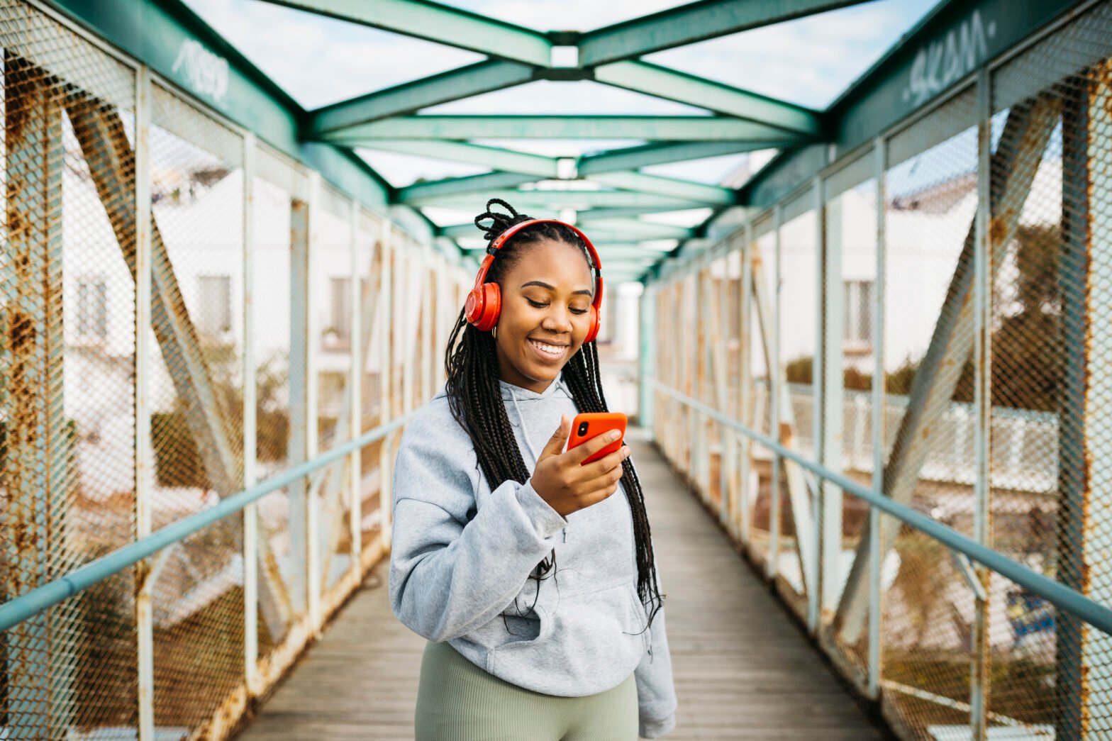 Portrait of a young smiling woman, with braided hair and sportswear, using a red smartphone and headphones, after workout on a urban bridge