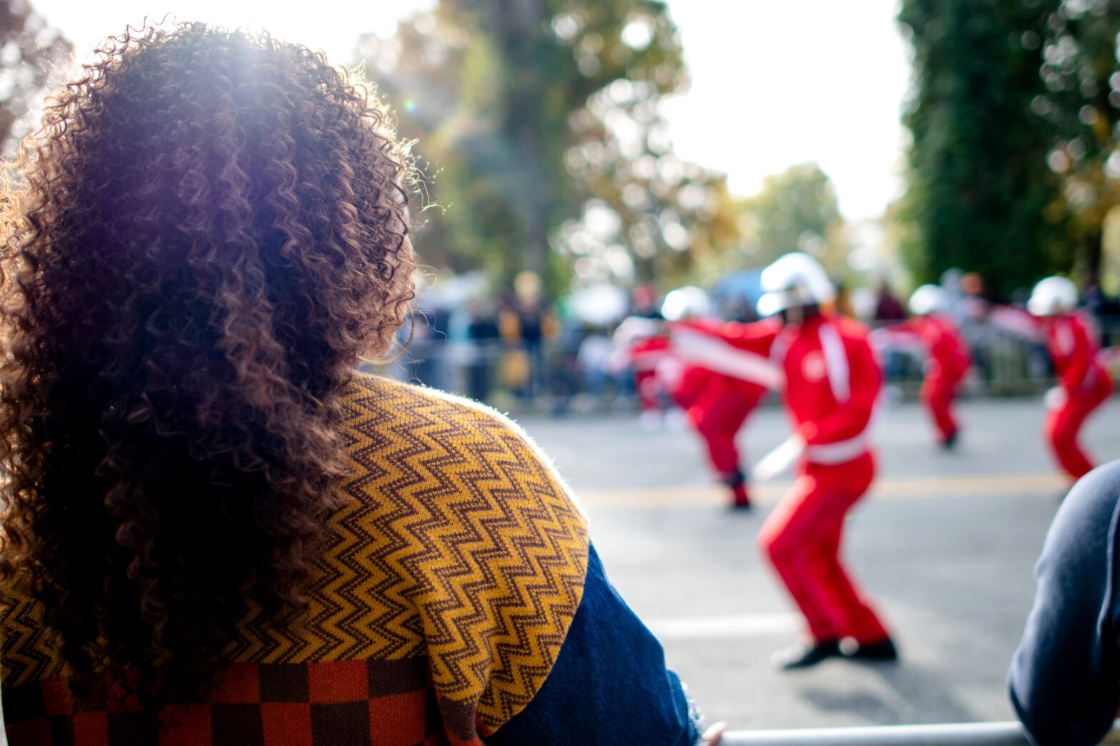 Black woman with curly hair watches a HBCU parade featuring dancers in red uniforms performing in the street, with spectators and trees blurred in the background.