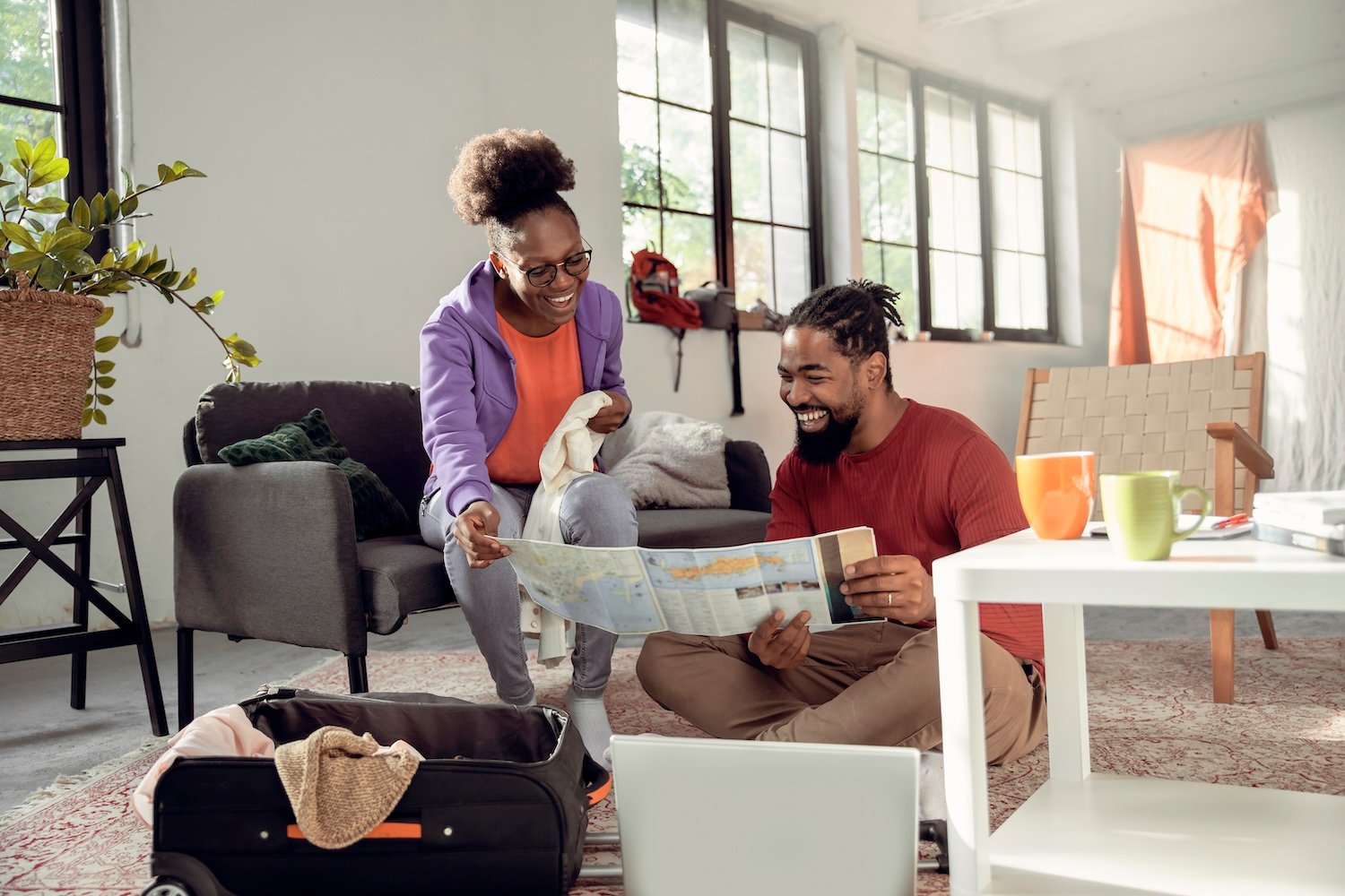 Smiling young couple looking at world map, planning vacation trip and packing suitcase in their living room.