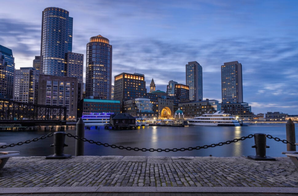 This view of the the Boston cityscape is particularly breathtaking due to the ocean's reflection of the skyscraper's radiant lights. This was taken in the blue hour, where the deep indigos of the sky perfectly complement the warmer glows of the city. The stone-paved road and iron chains are an iconic signifier of this district, especially as these historically lasting features contrast the modern skyscrapers.