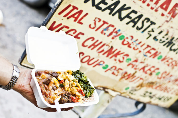 Kingston, Jamaica - January 19, 2012: Hand holding a take away plate of vegetarian rasta food (ital food) in front of the restaurant sign in downtown market area.