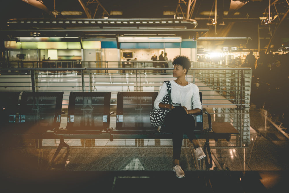 Beautiful young Black female tourist is sitting inside of modern airport terminal and waiting for her flight; cute Brazilian girl with curly Afro hair is sitting in waiting hall of railway station