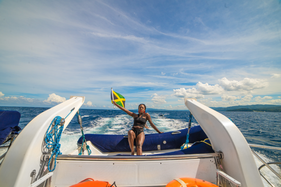 A person in a black swimsuit waving a Jamaican flag poses at the back of a boat on the ocean. 