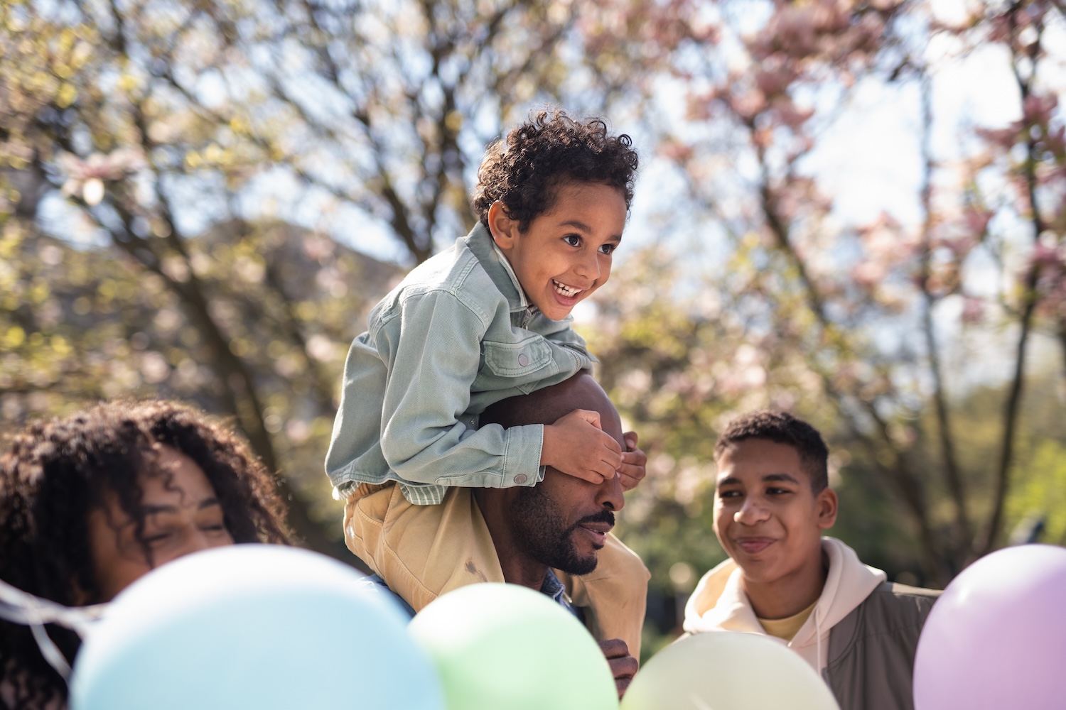 Family enjoying a sunny spring day with pastel balloons and blooming trees in the background, capturing the joy of Easter celebrations.