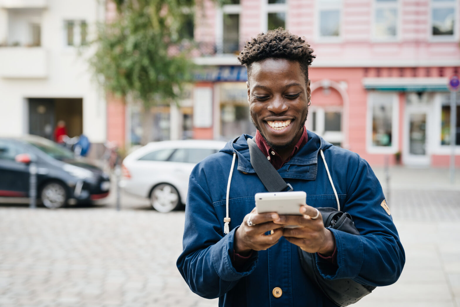 A man smiling while using his smartphone to message friends while out in the city.