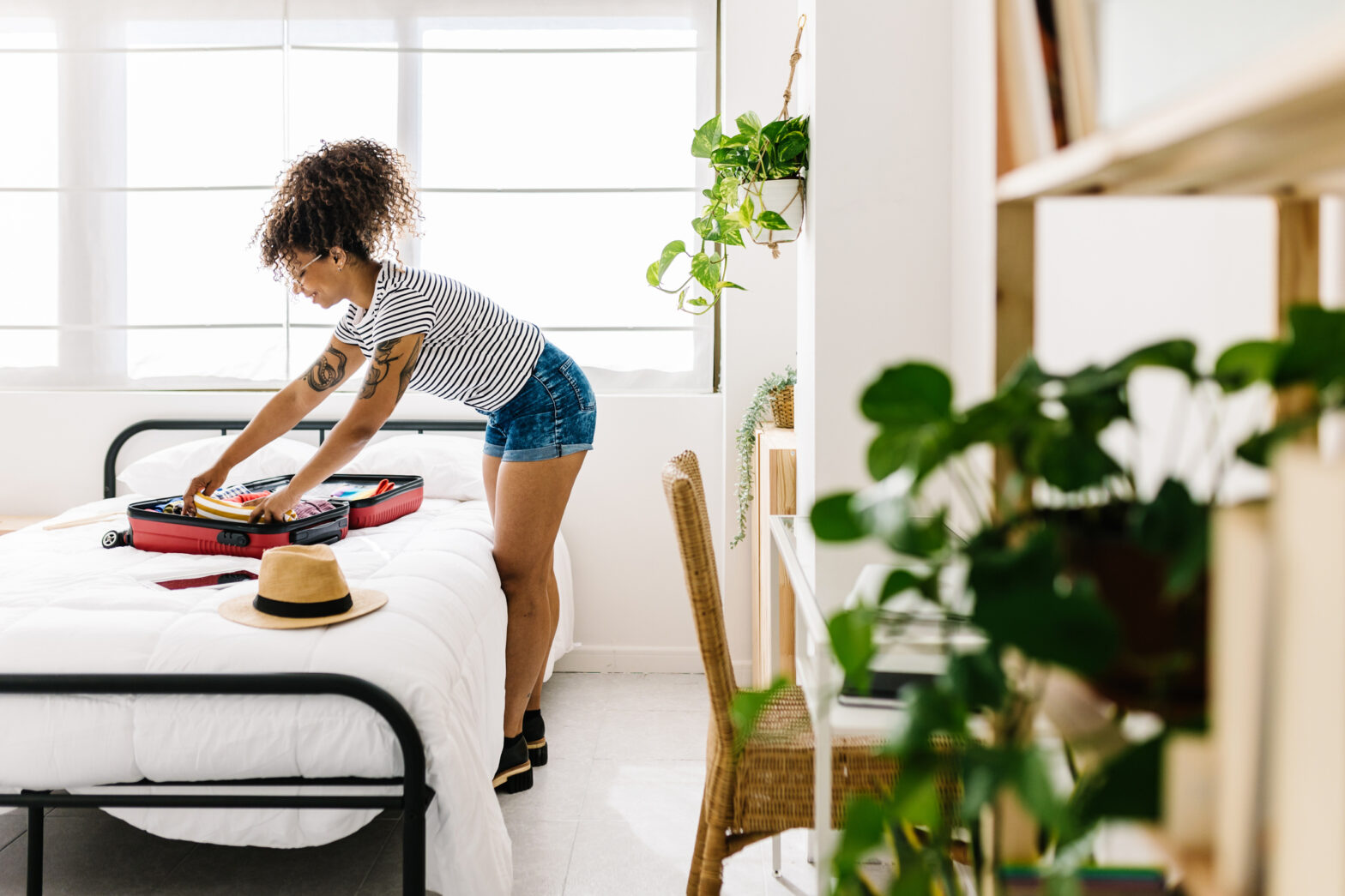 Young woman packing clothes into her travel suitcase on the bed - Afro american female getting ready for summer vacations preparing luggage for a journey trip