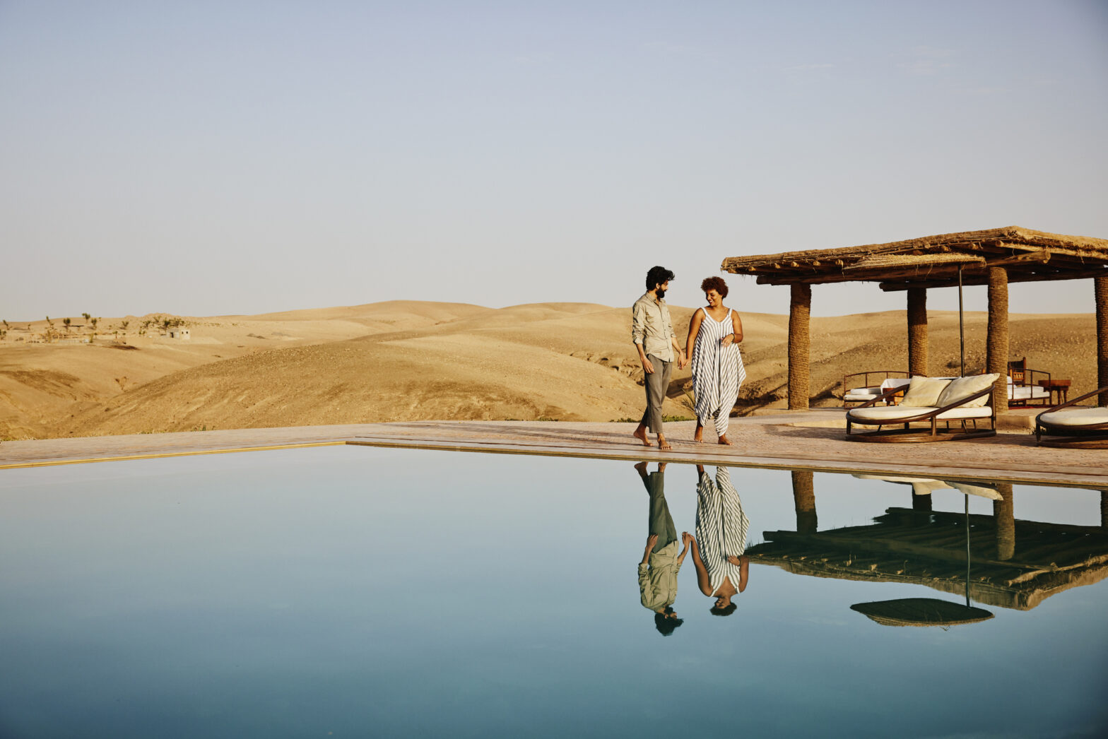 2023 Travel Noire Awards: Favorite Unique Stay. Wide shot of couple walking around pool at Moroccan desert camp at sunrise while on vacation