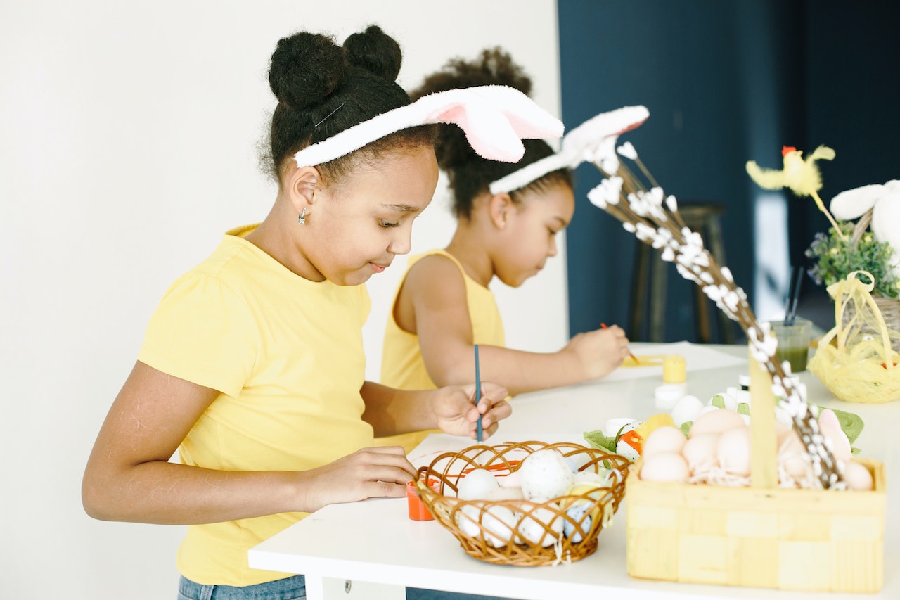 Get inspiration from around the world on what to name your twin girls with this list of twin girl names. Pictured: Two little girls wearing matching yellow shirts and bunny ears painting.