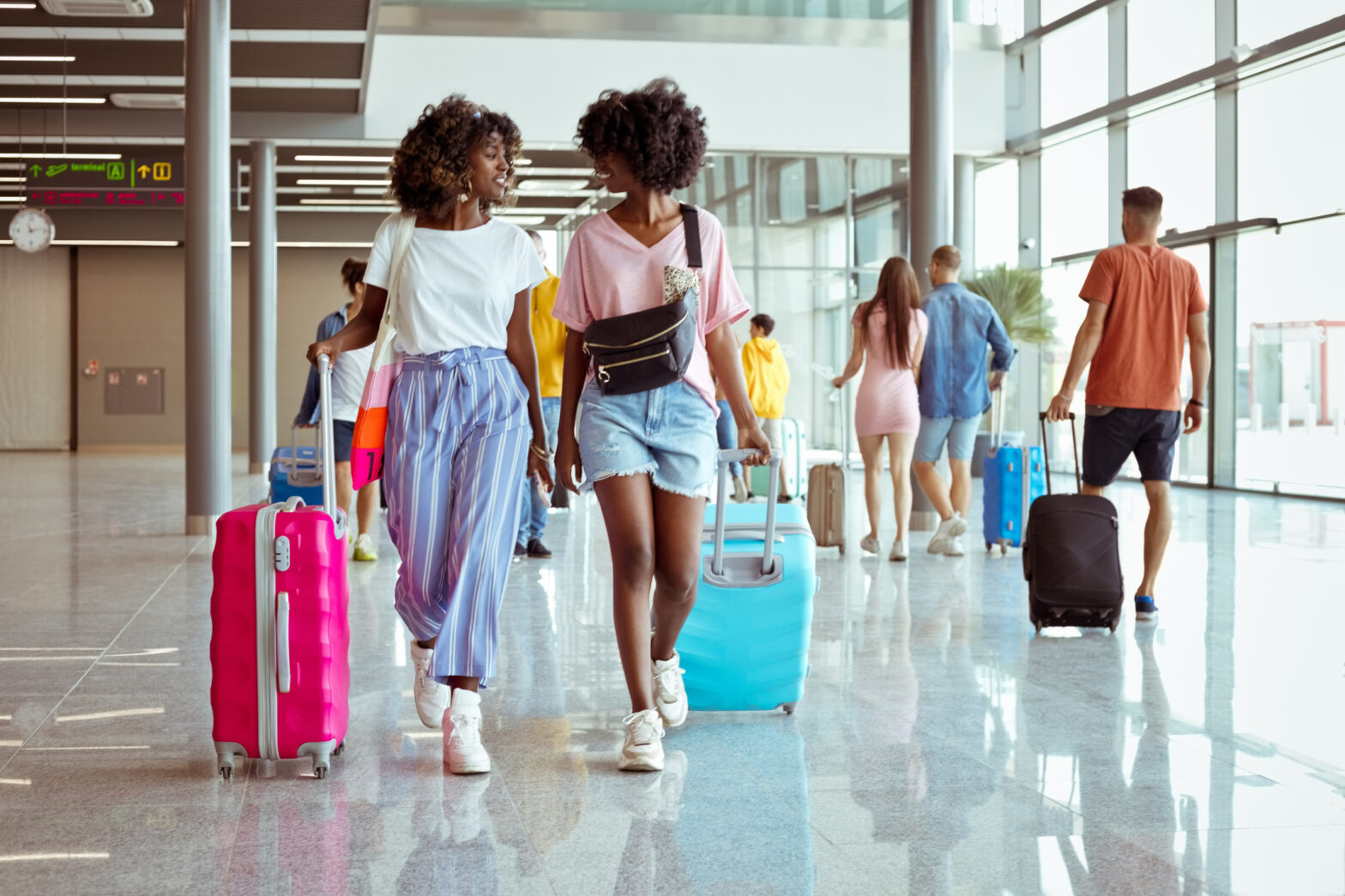 Passengers walking with wheeled luggage at airport terminal. Two young women on foreground.