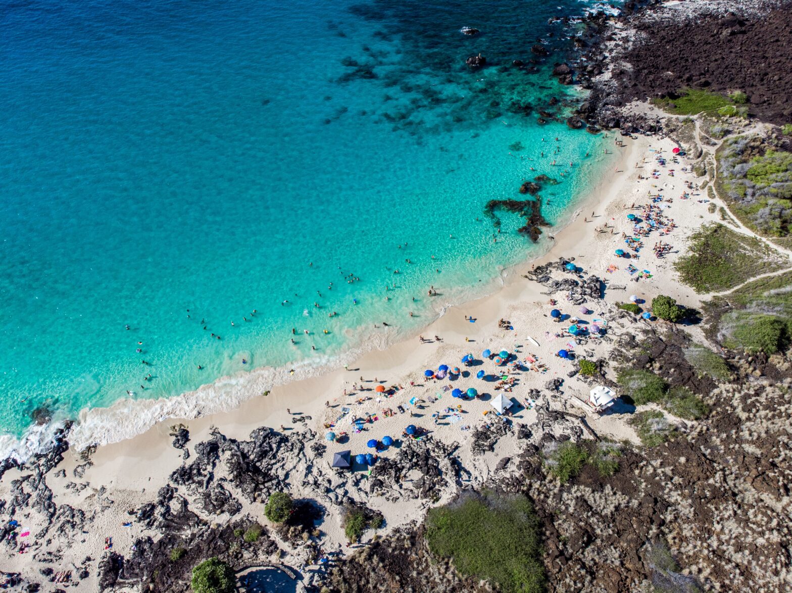 The Magic Sands Beach park is a sunny and popular place for tourists in Hawaii, the Big Island. Pictured: The Kona Beach filled with beachgoers and black sandy rocks.