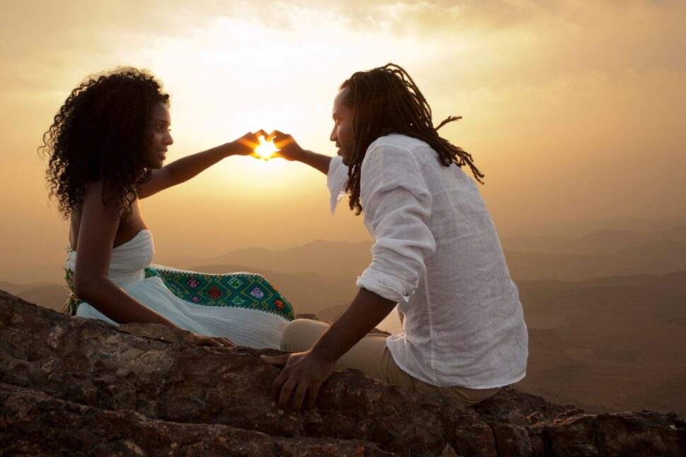 couple sits in front of sunset and make heart shape with hands