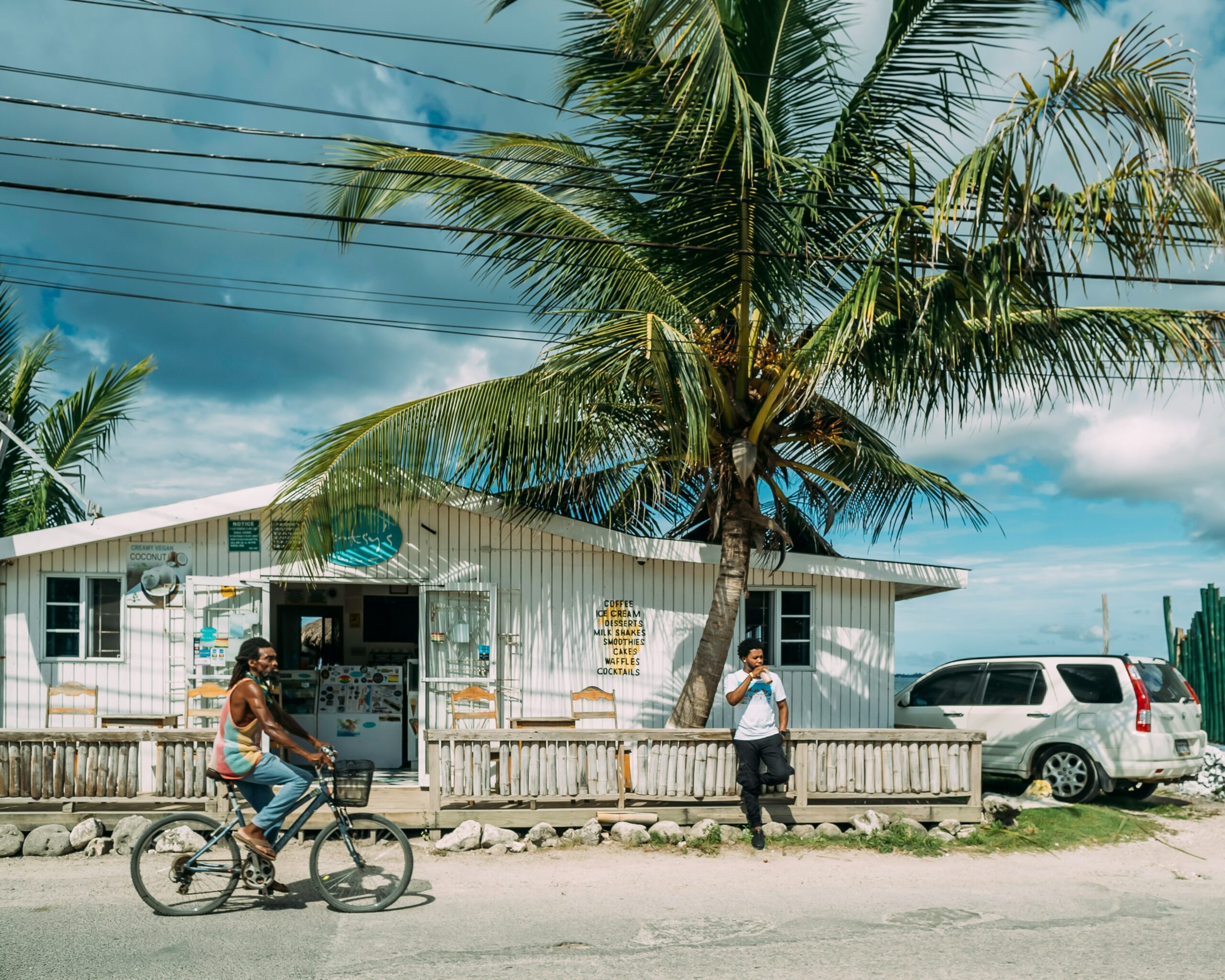 Jamaica is a popular tourist destination but travelers often wonder if it is safe. Learn more about Jamaica’s safety situation.
Pictured: a white one-leveled building with a large palm tree in front of it with local Jamaicans in front of it