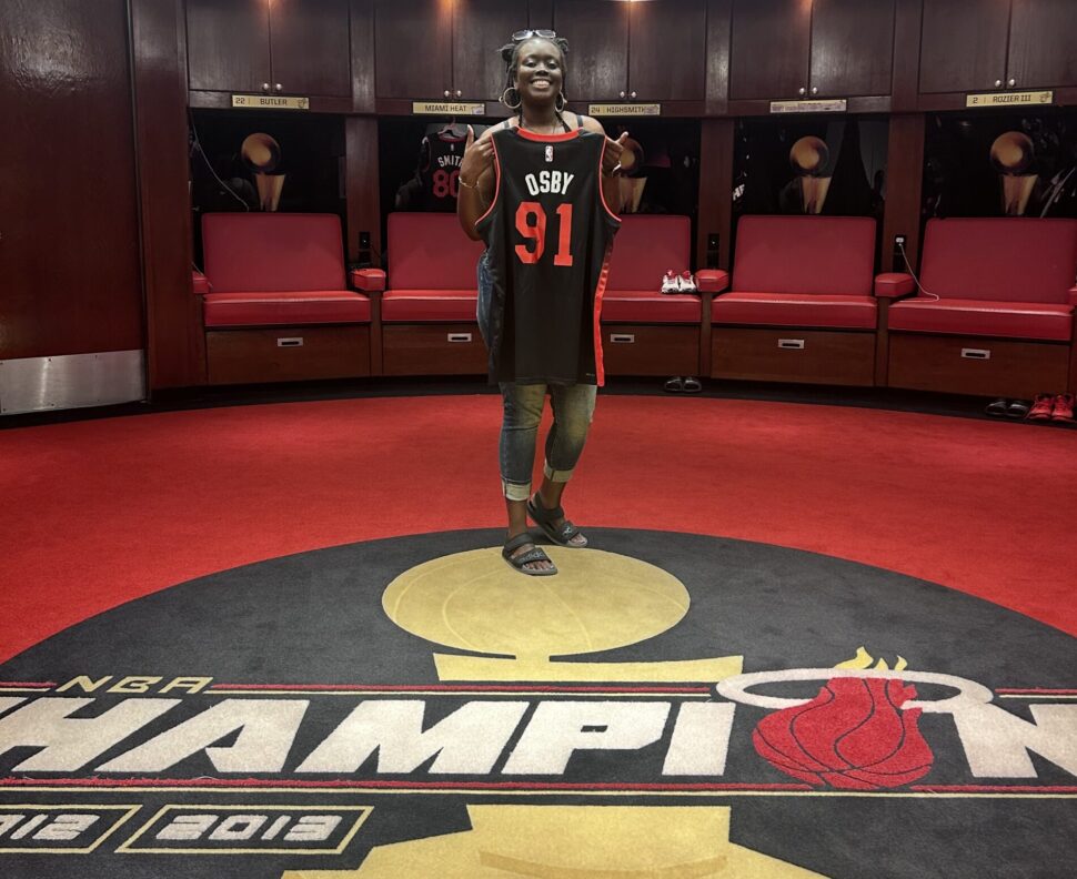 Osby standing with her custom Miami Heat jersey in the home team locker room at the Kaseya Center. 