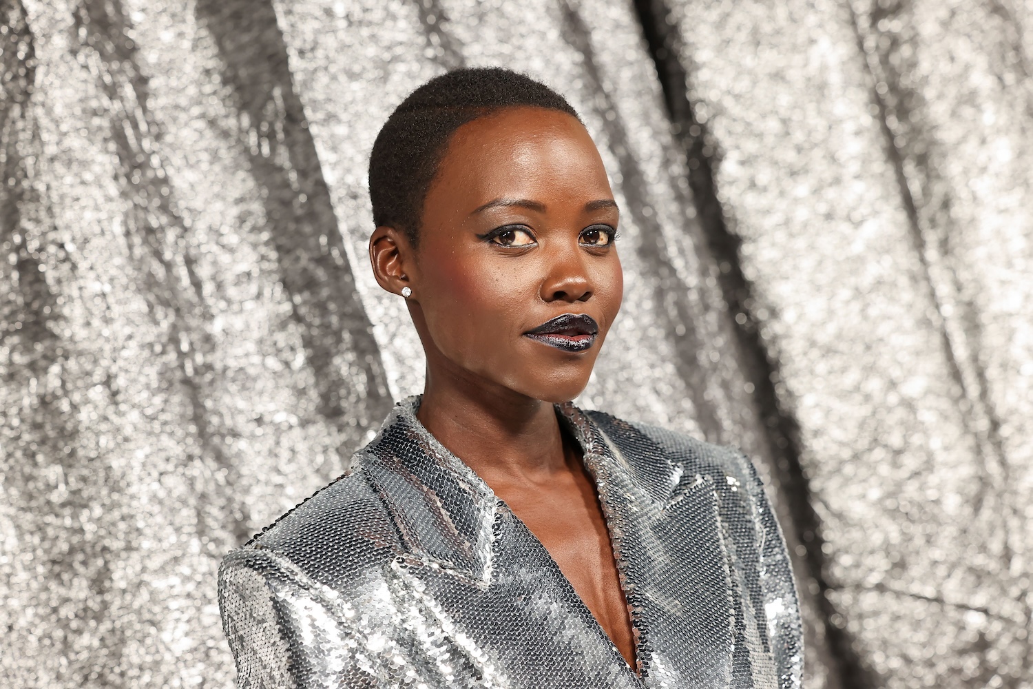 Lupita Nyong'o wearing a shimmering silver outfit against a silver backdrop, looking confidently at the camera.