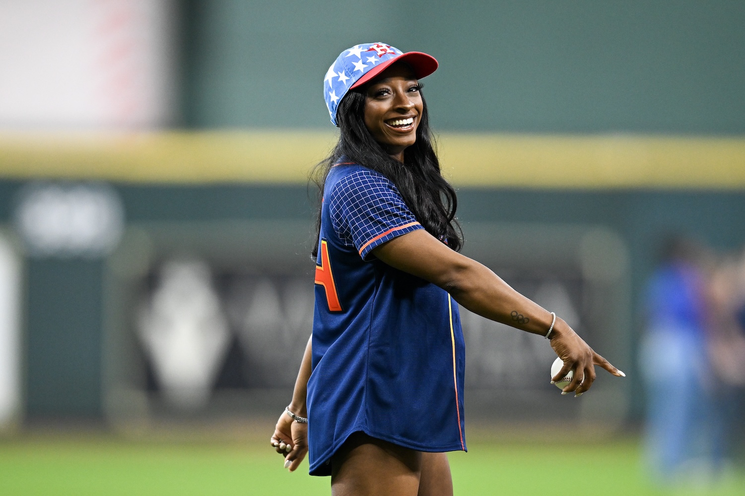 Simone Biles, smiling and wearing a Houston Astros jersey and a blue baseball cap with stars, prepares to throw a baseball on the field at an Astros game.