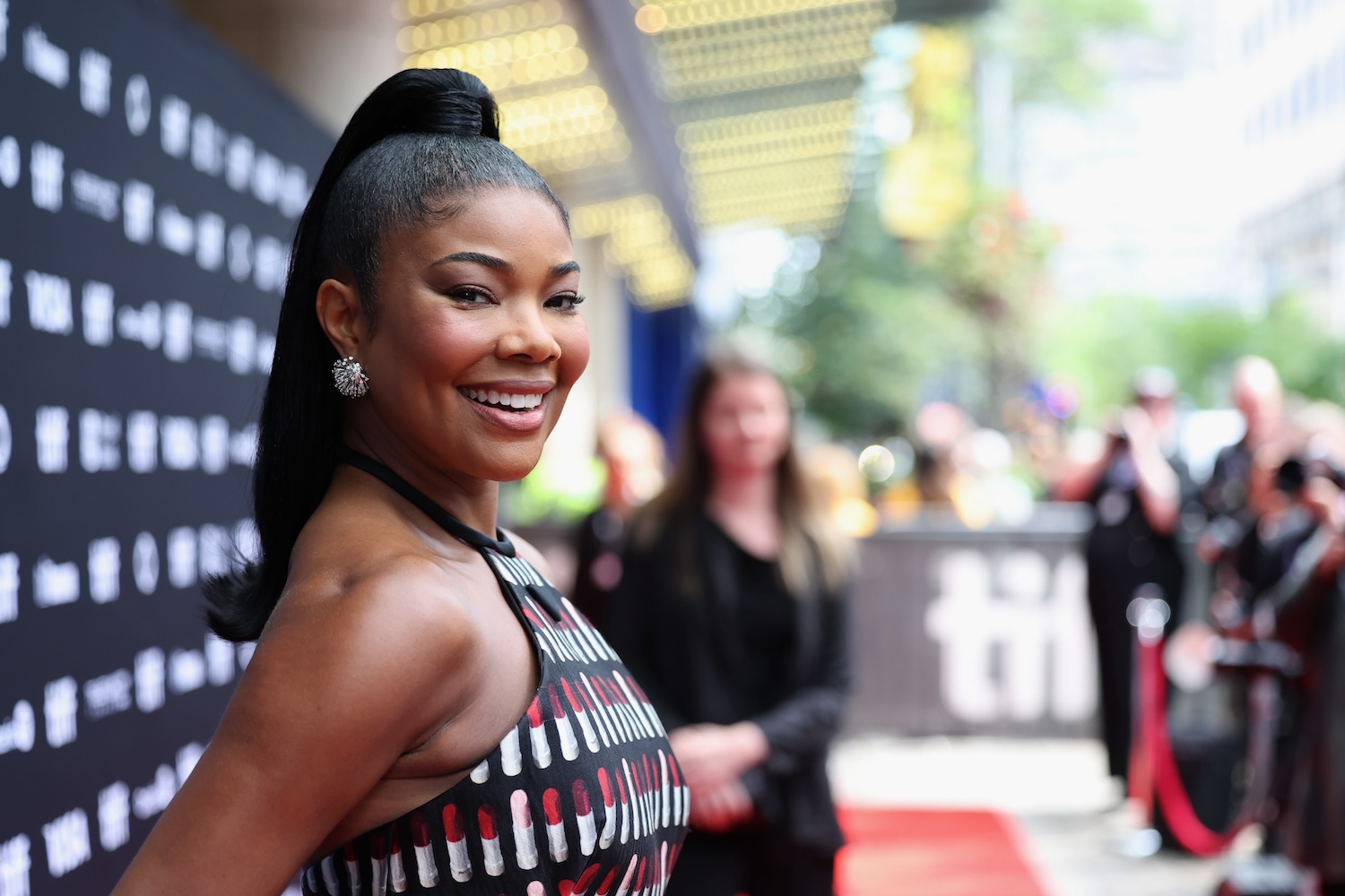 Gabrielle Union smiles on the red carpet, wearing a black halter dress with lipstick prints and a sleek high ponytail, with a blurred crowd and lights in the background.