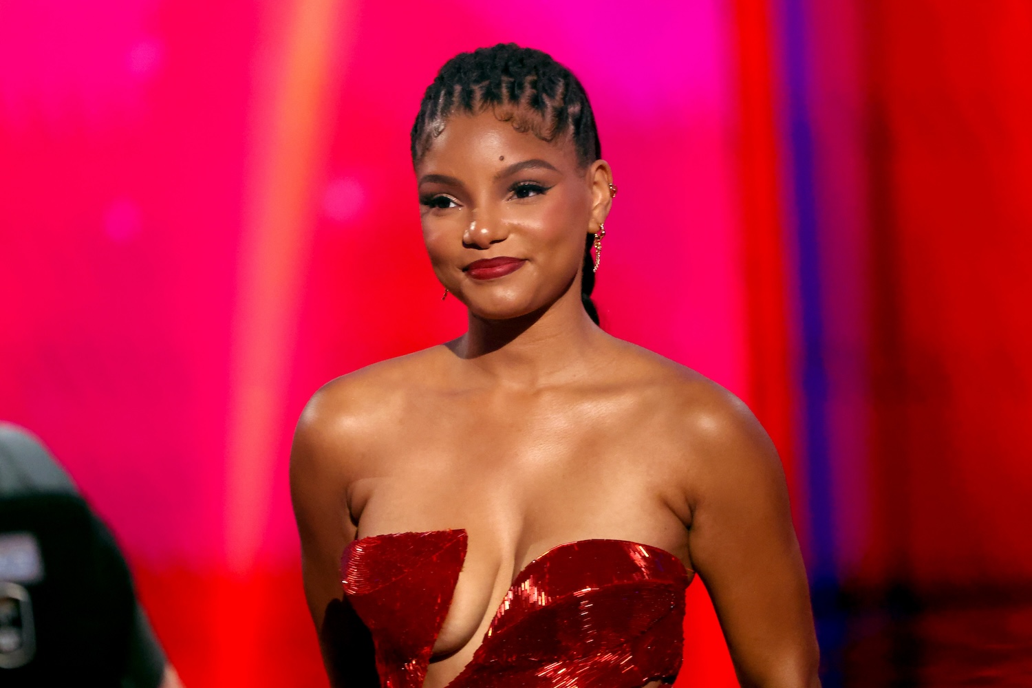 Halle Bailey is seen wearing a strapless red dress with a plunging neckline and her hair styled in neat cornrows, smiling against a vibrant pink and red backdrop.