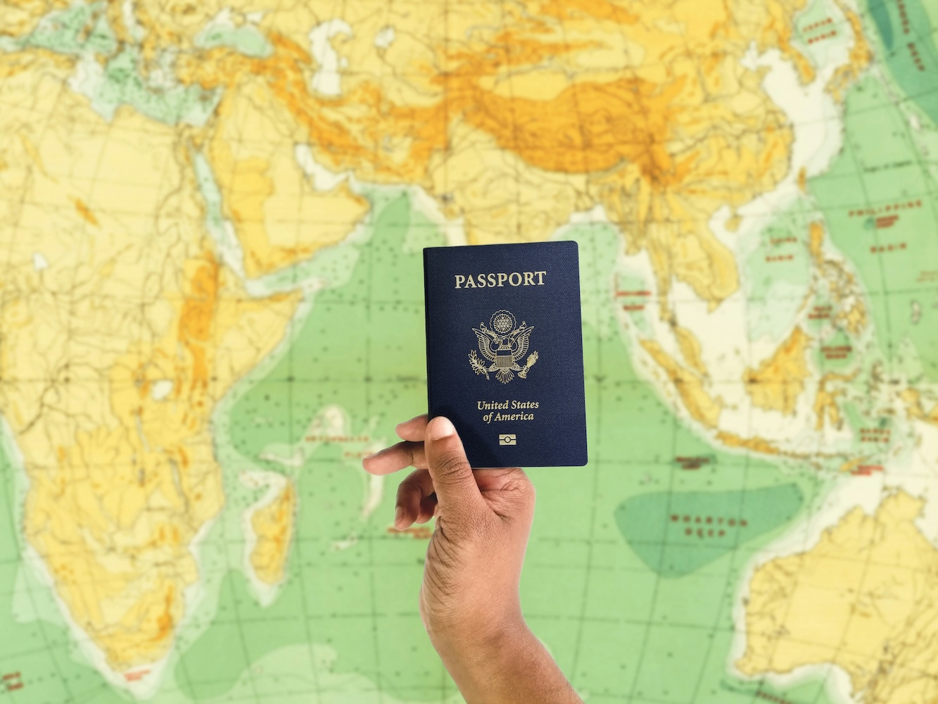 Hand holding a passport in front of a yellow and green world map.