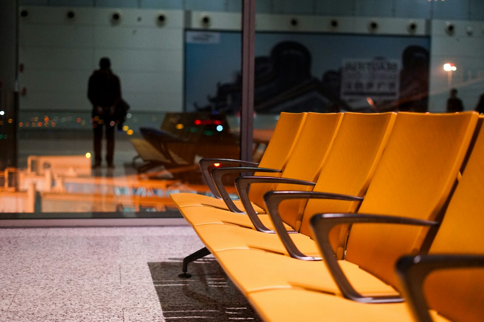 mpty orange airport seats in a waiting area, with a blurred figure in the background standing near large glass windows overlooking the runway at night.