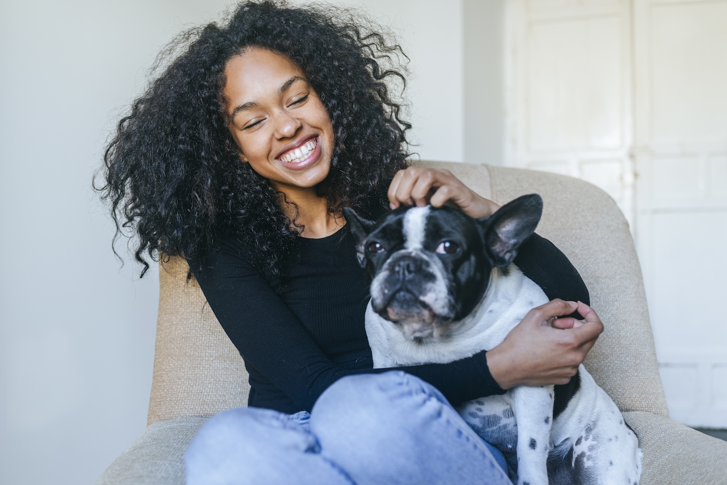 A young woman with curly black hair sits on a beige armchair, smiling joyfully while cuddling a black and white French Bulldog.