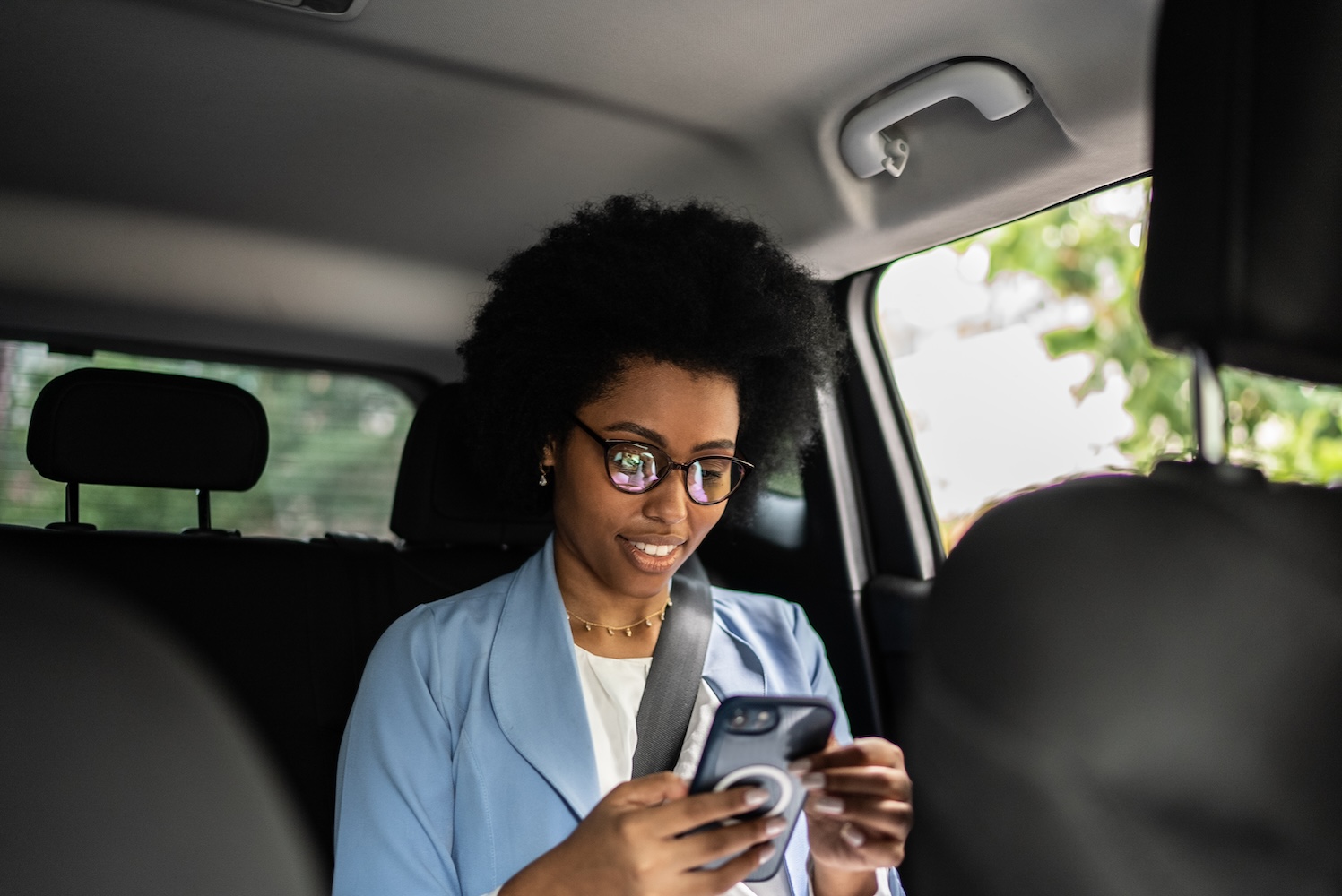 A businesswoman in a light blue blazer sits in the backseat of a black car, focused on her smartphone while wearing glasses and a seatbelt.