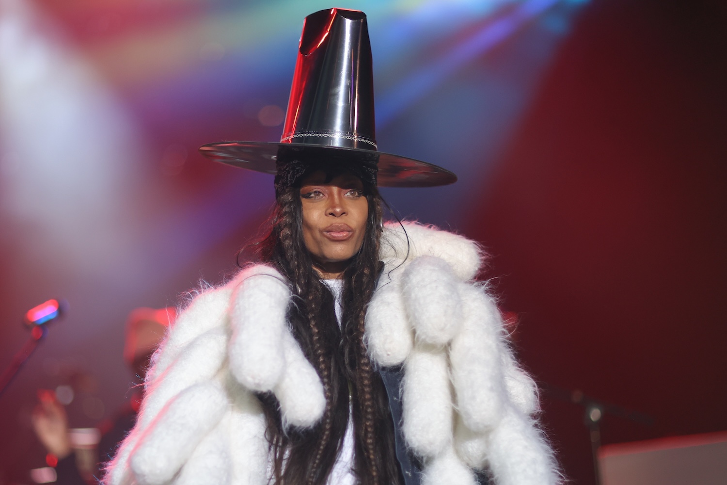 Erykah Badu performing on stage wearing a large, metallic, cone-shaped hat and a voluminous white fur coat with braided hair, under colorful stage lights.