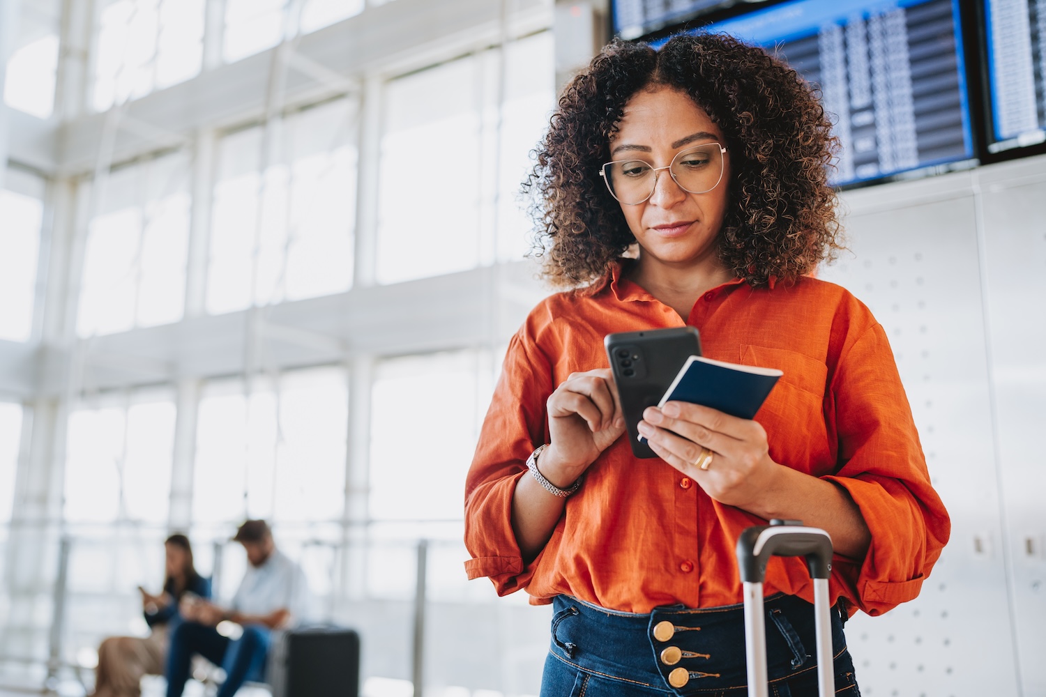 An airport traveler in an orange shirt checks her phone while holding a passport, with luggage beside her and flight information screens in the background.