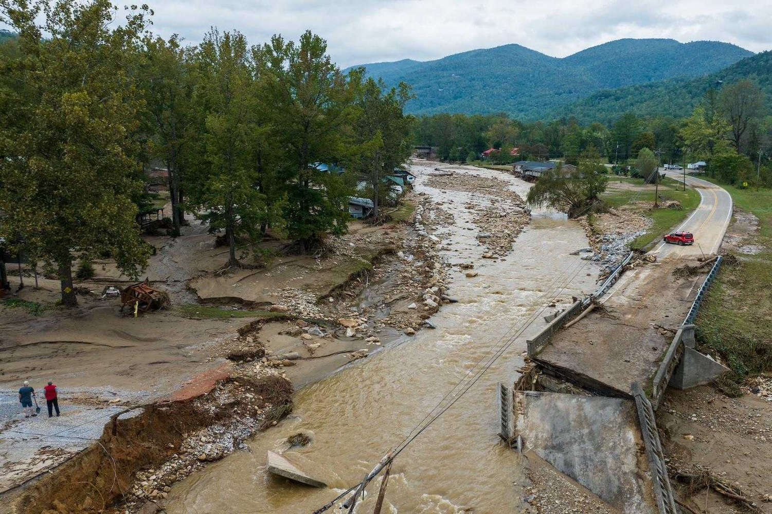 A collapsed bridge and flood-damaged road by a debris-filled river, with two people surveying the destruction in a rural, mountainous area.