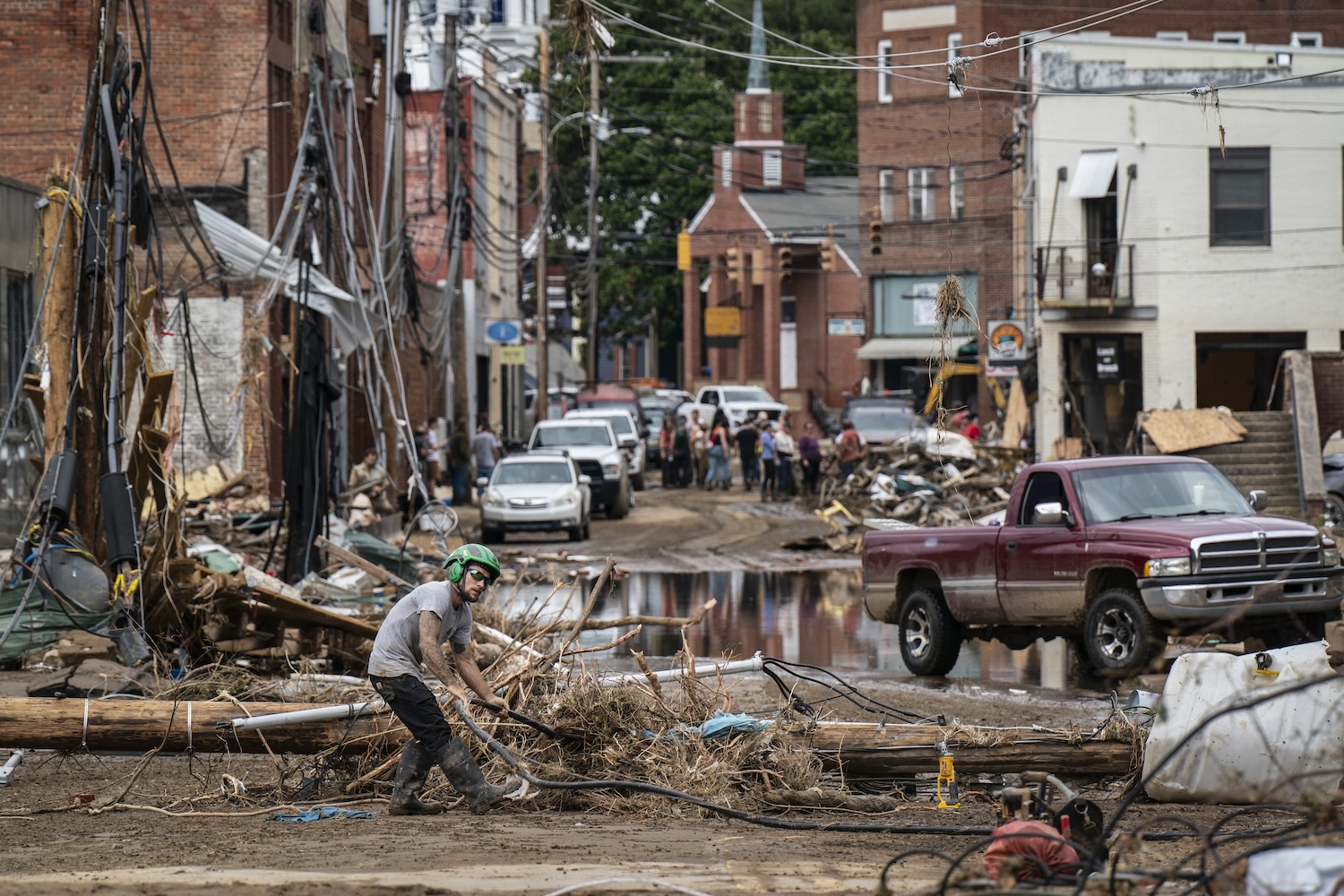 A worker clears debris from a flooded street in Marshall, North Carolina, damaged by Hurricane Helene, surrounded by downed power lines, damaged buildings, and wreckage.