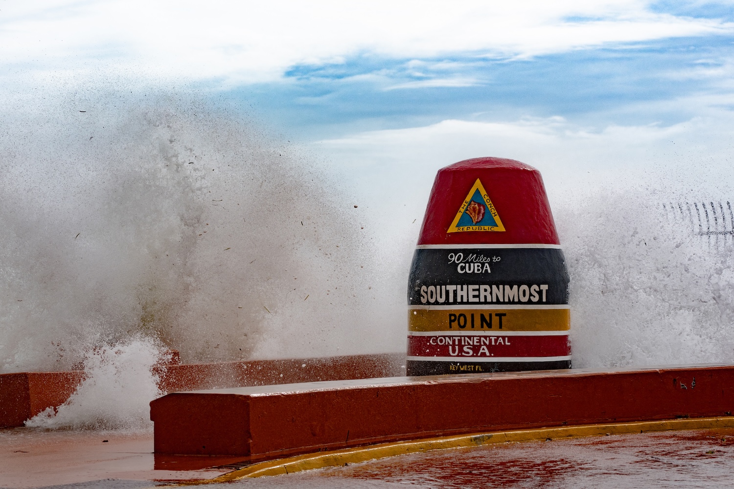 A large wave crashes against the Southernmost Point buoy in Key West, Florida, marked with the inscription "90 Miles to Cuba" and "Southernmost Point Continental U.S.A." The water splashes over the buoy as stormy weather stirs up the sea under a partly cloudy sky.