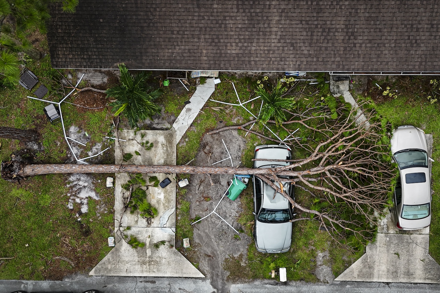 An aerial view shows a large tree toppled over onto two parked cars in front of a house after Hurricane Milton damage, with debris scattered around the property.