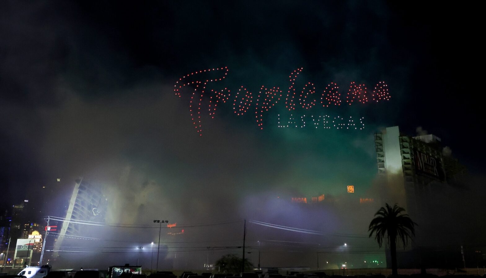 The image captures the Tropicana Las Vegas demolition at night, with dust filling the air and "Tropicana Las Vegas" illuminated above in lights as the building collapses.