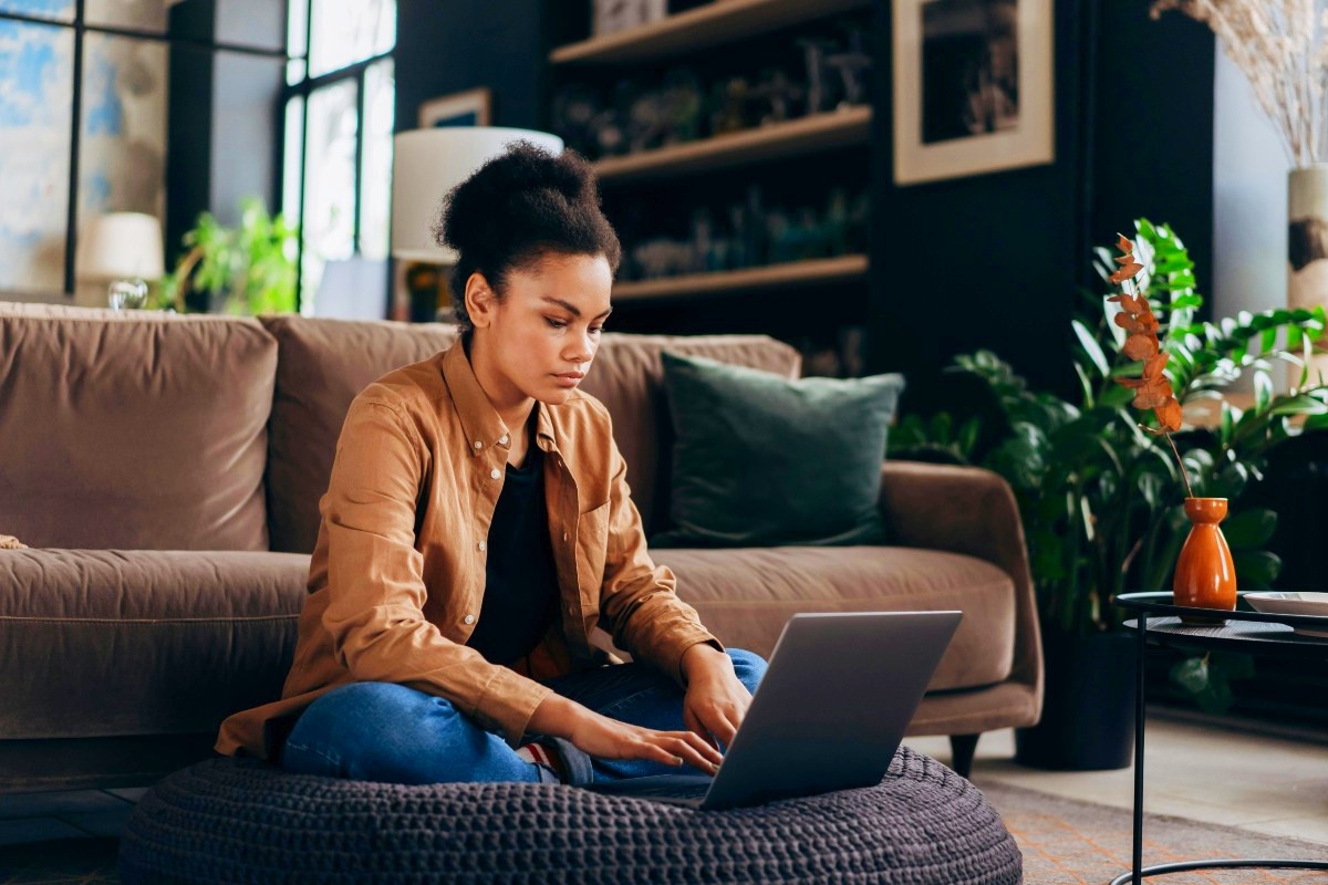 Woman sitting on black knitted floor cushion while working on her laptop.