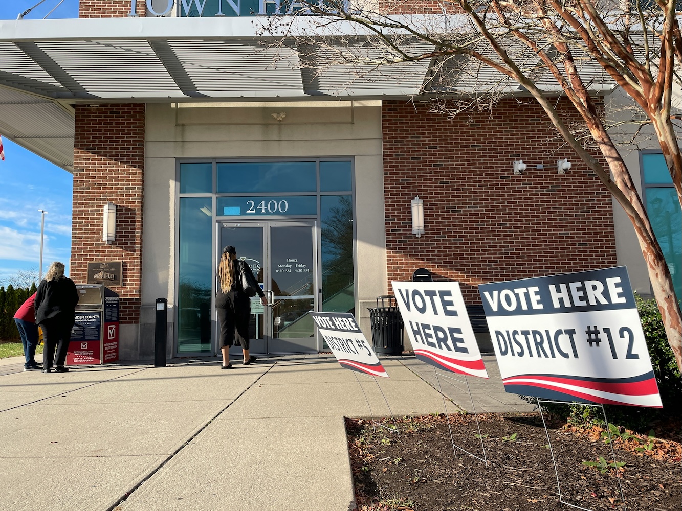 People entering a town hall building marked as a voting location, with 'Vote Here' signs indicating different districts in the foreground.