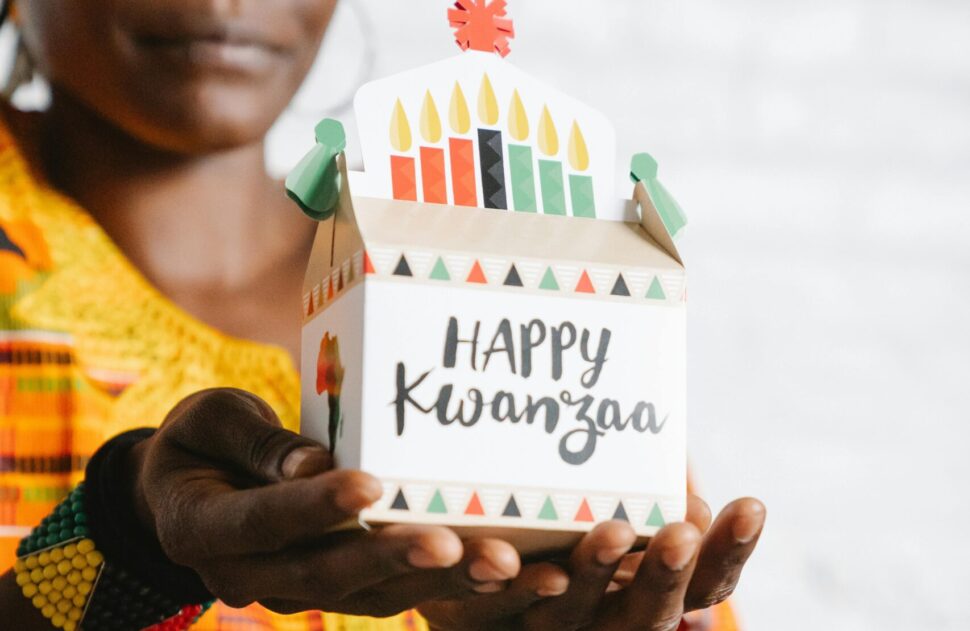 A woman holding a small gift box with "Happy Kwanzaa" written on the side.