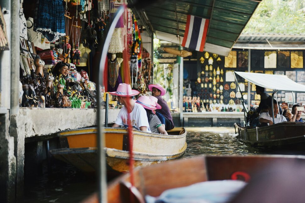 A boat cruising through Bangkok's Damnoen Saduak Floating Market. People in pink hats look at figures of cats and other animals from the boat. 