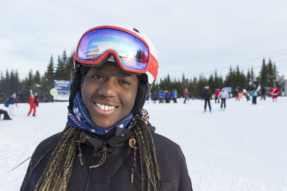 A smiling Black skier wearing a red helmet and reflective goggles stands on a snowy slope, surrounded by other skiers under an overcast sky.