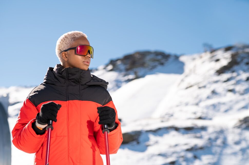 A Black skier with platinum hair and reflective goggles stands confidently on snowy slopes under a clear blue sky.