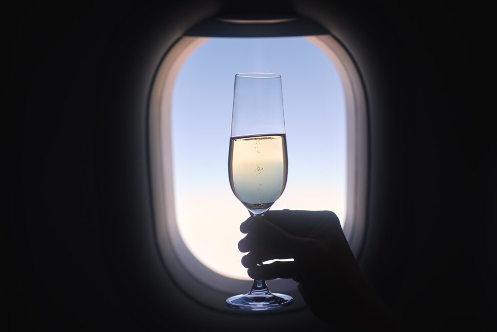 Close-up of silhouette hand holding champagne glass against airplane window during flight on sunny day.