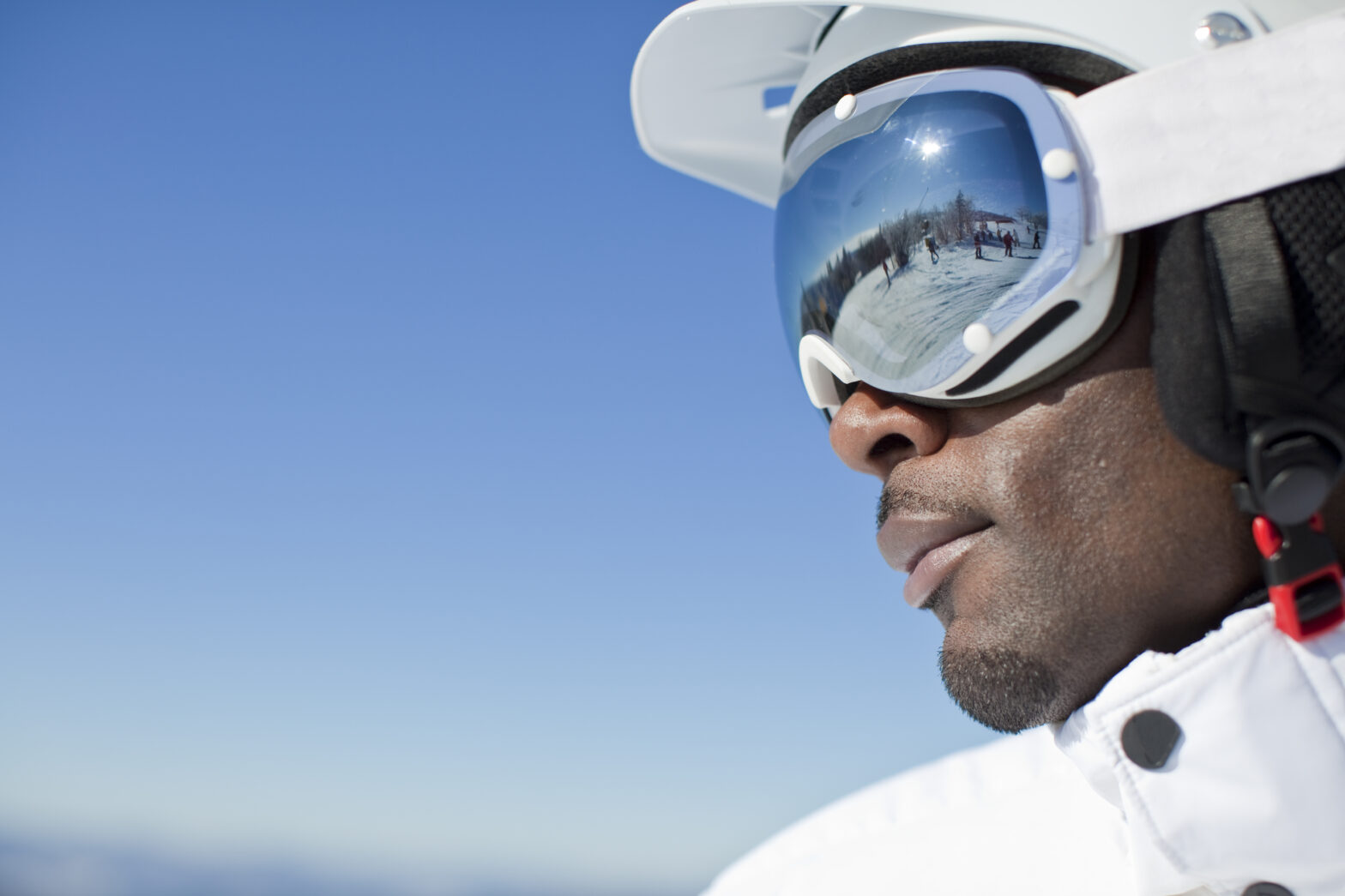 A Black skier wearing reflective goggles and a white helmet gazes confidently toward a snow-covered mountain under a clear blue sky.