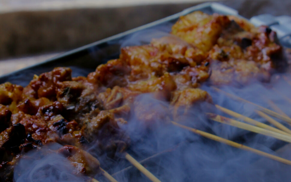 Close-up of skewered, grilled meat coated in sauce, with smoke wafting over the sizzling barbecue.