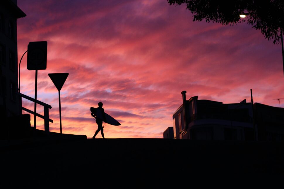 Australian surfer at dawn walking under pink and orange fiery hues in the sky.