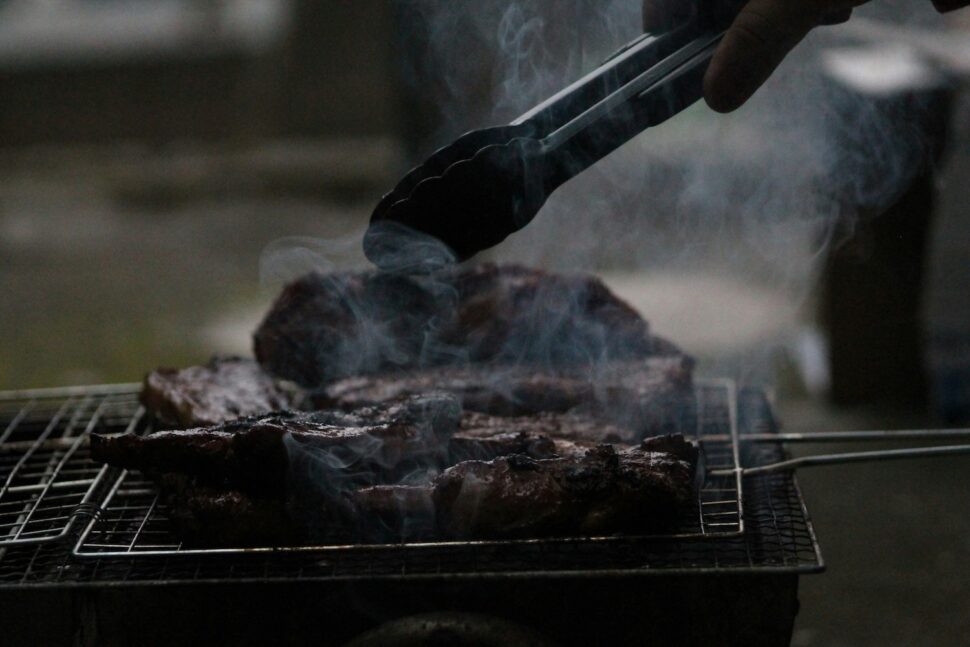 Close-up of a barbecue grill with sizzling meat, smoke rising as tongs are used to flip or adjust the pieces.