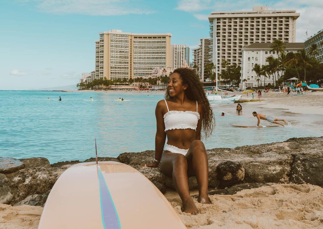 Woman sits next to her surfboard with Honolulu's beach area in the background