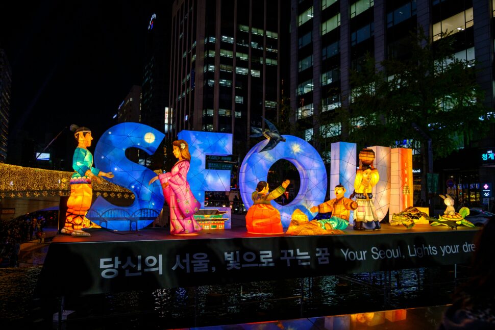 The word 'Seoul' illuminated with paper figures, set against a backdrop of modern skyscrapers of Seoul, South Korea.