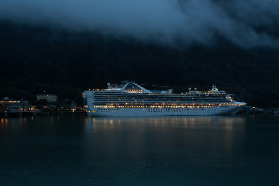 A cruise ship docked at a bustling port at night, glowing with warm lights that reflect off the calm water.